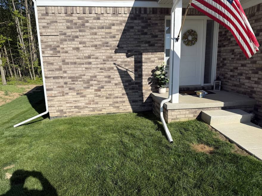Front corner of a brick house in Canton, Michigan with two white downspout extensions lying on the lawn, emptying roof water a few feet from the foundation.